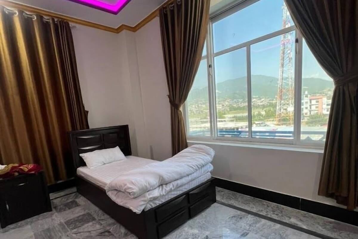 Un dormitorio con cama, mesita de noche, ventana con cortinas y vistas al exterior.