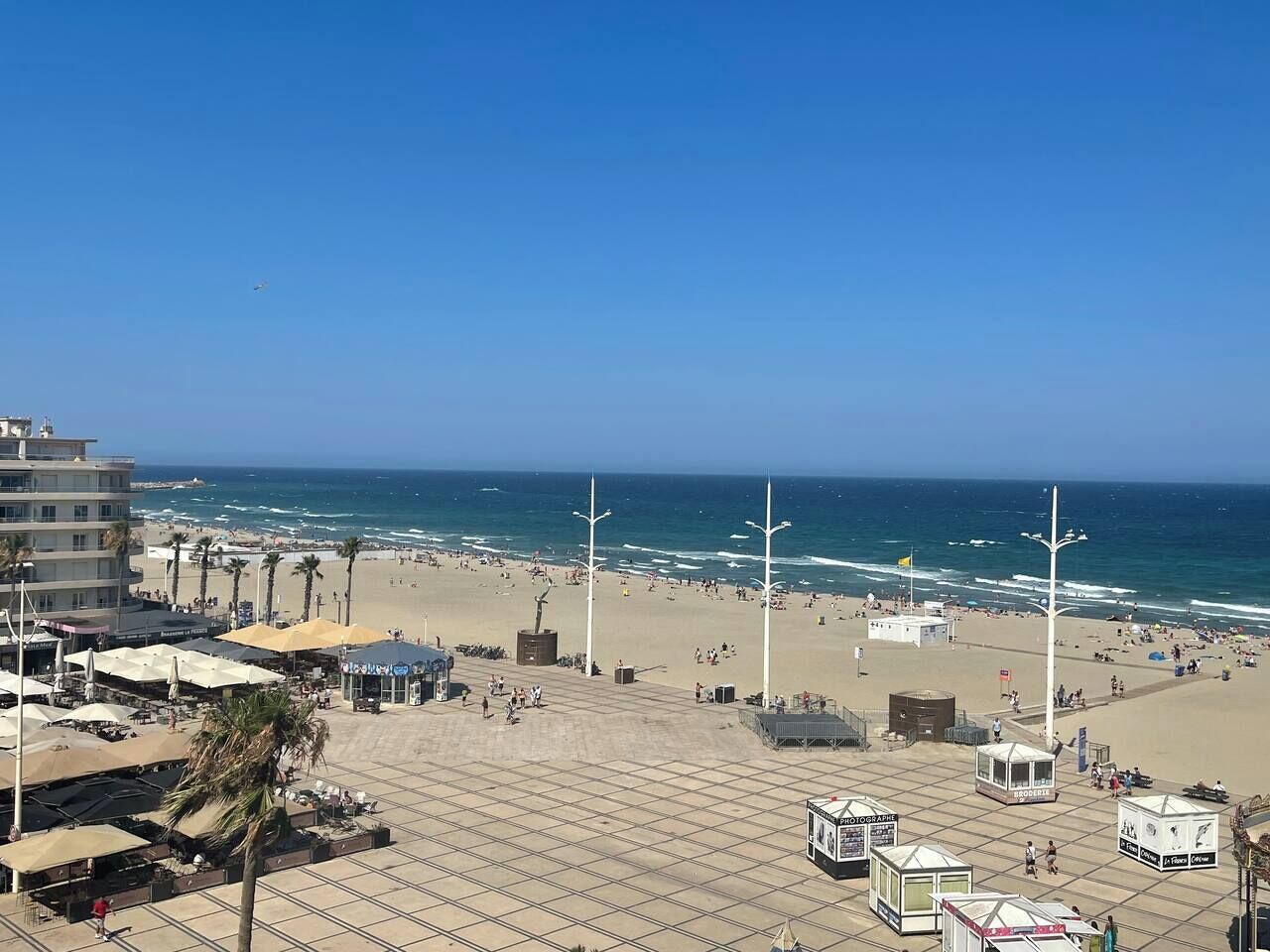 A beachside promenade with a view of the ocean, palm trees, and beach umbrellas.