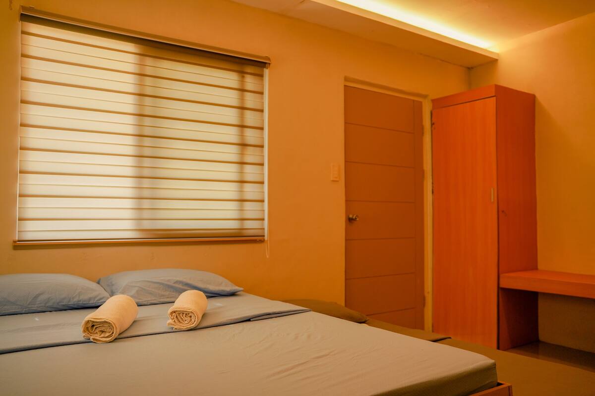 A single bed with a blue mattress and pillows, a wooden wardrobe, and a window with blinds.