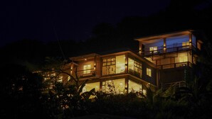 Una casa moderna de dos pisos con varios balcones y ventanas amplias, rodeada de vegetación densa.
