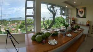 Una cocina con vista a un paisaje tropical, un trípode y una mesa de comedor dispuesta con platos y una bebida.