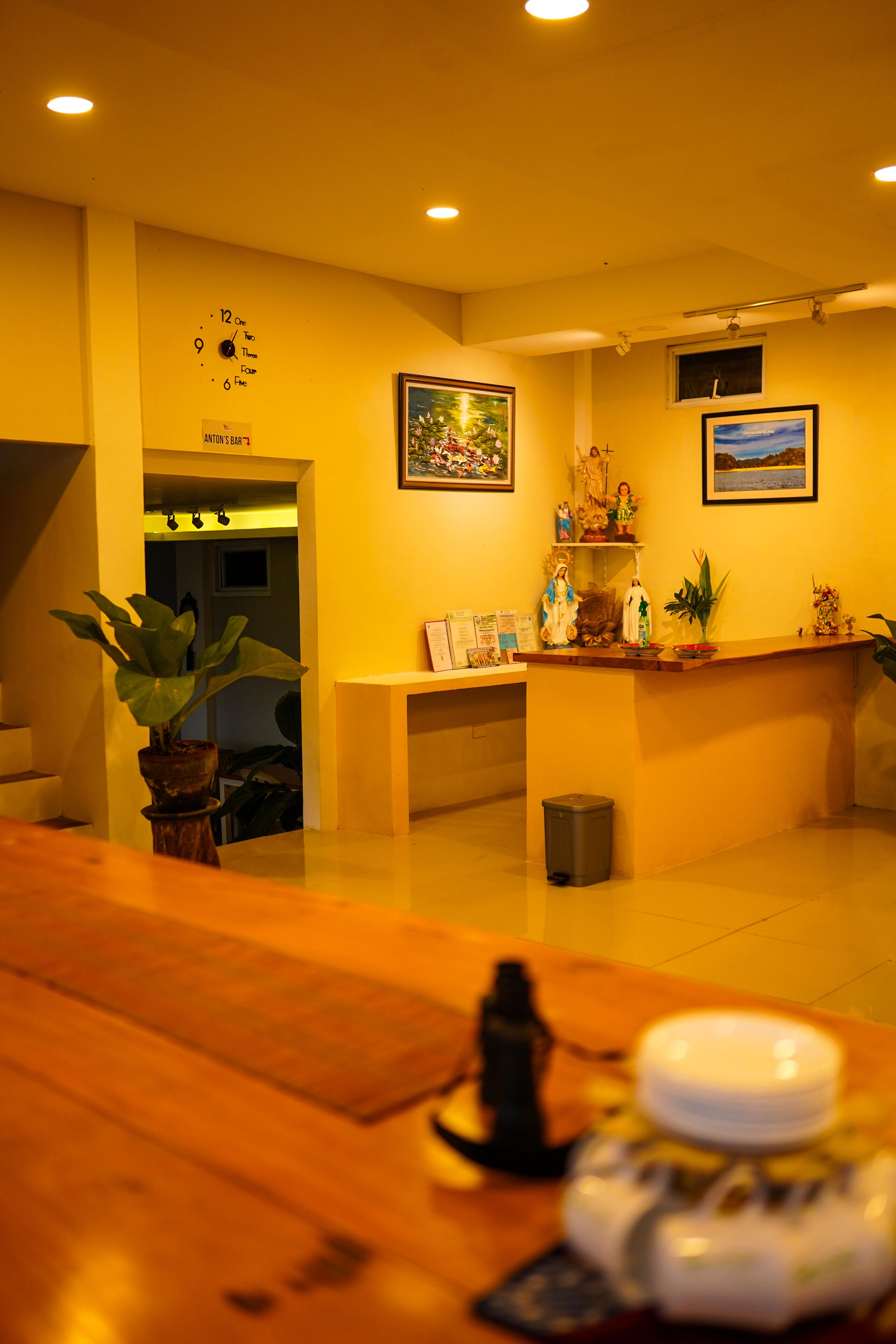 Uma área de recepção de hotel com um balcão de madeira, um relógio, um quadro emoldurado e uma pequena lixeira.