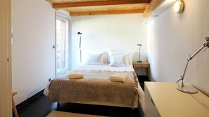 Une chambre avec un plafond en bois, un lit recouvert d’une couverture beige, une lampe de chevet et une table de nuit.