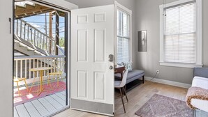 A room with a sliding glass door leading to an outdoor deck, a bed with a white comforter, a window with blinds, and a small table with chairs.