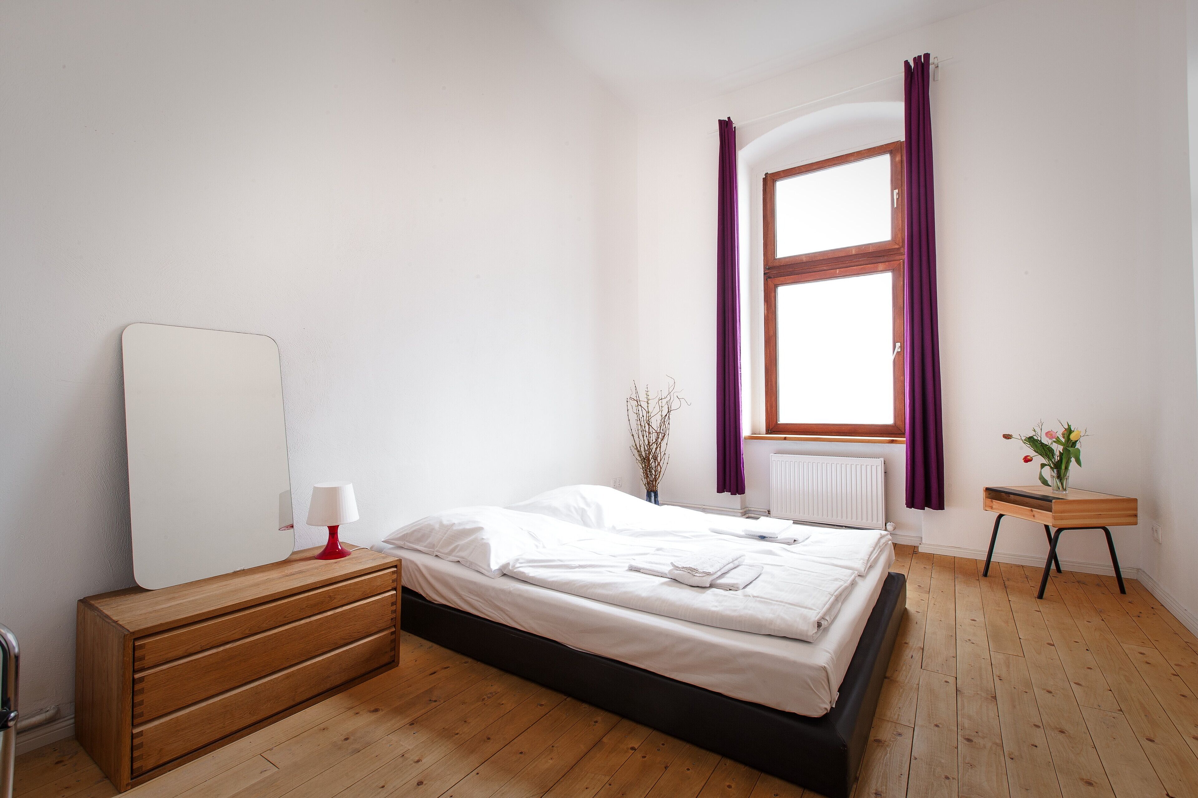 A bedroom with a wooden bed, a dresser, a mirror, a window with purple curtains, and a small table with a vase.