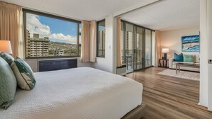 Hotellihuone, jossa on suuri sänky, näkymä rakennuksiin, liukuovi parvekkeelle ja taulu seinällä.