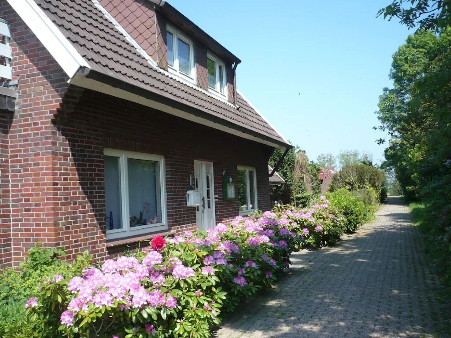 A brick house with a sloped roof, a front door, and a window. There is a pathway leading to the house surrounded by flowering bushes.