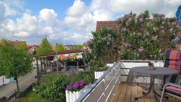 A wooden deck with a table and chairs, surrounded by lush greenery and purple flowers.