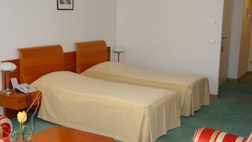 A hotel room with two single beds, a wooden nightstand with a lamp, a small table with a vase of flowers, and a striped armchair.