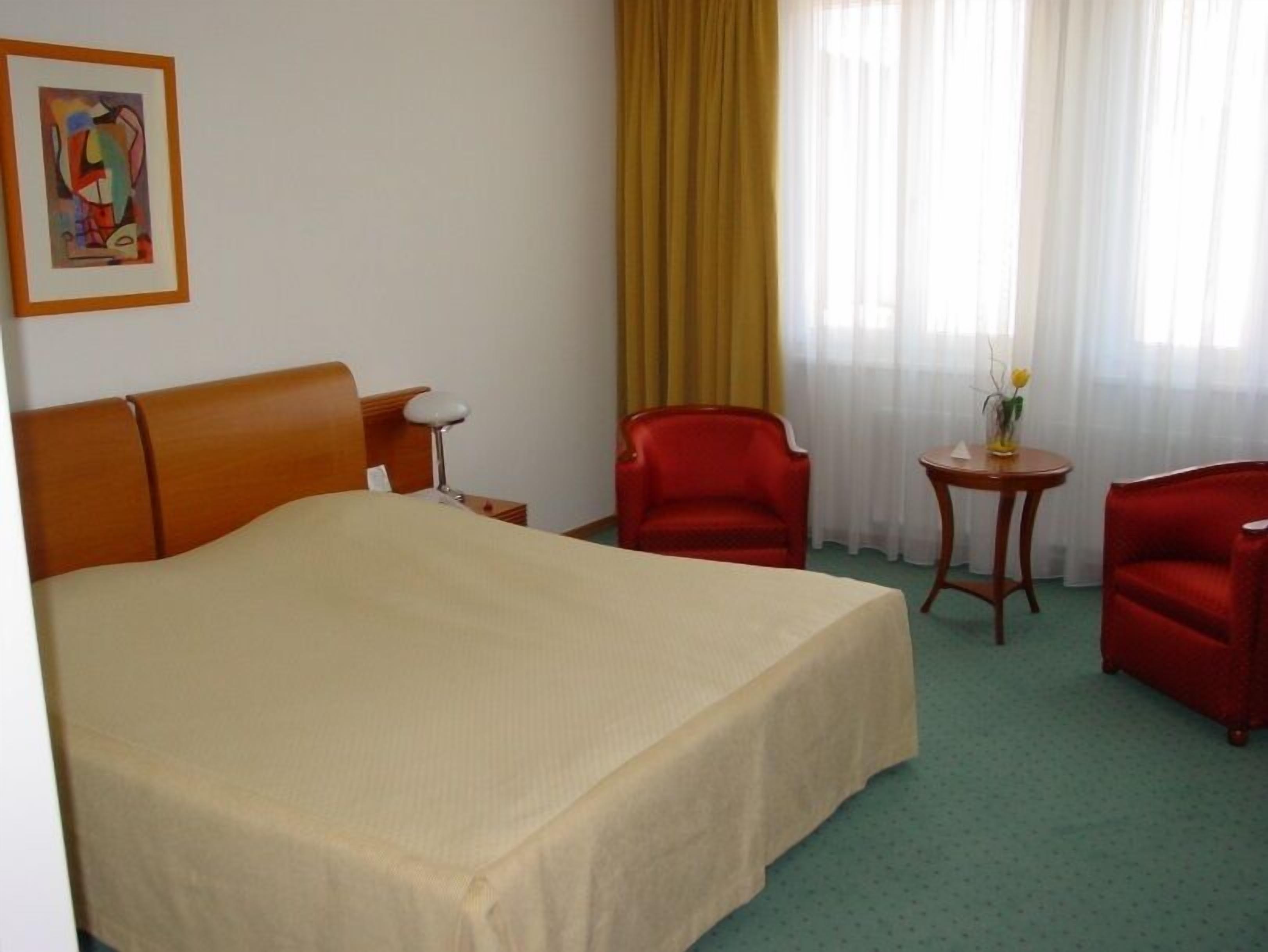 Una camera d'albergo con un letto grande, una poltrona rossa, una seconda poltrona più piccola, un tavolino con un vaso e un quadro appeso al muro.