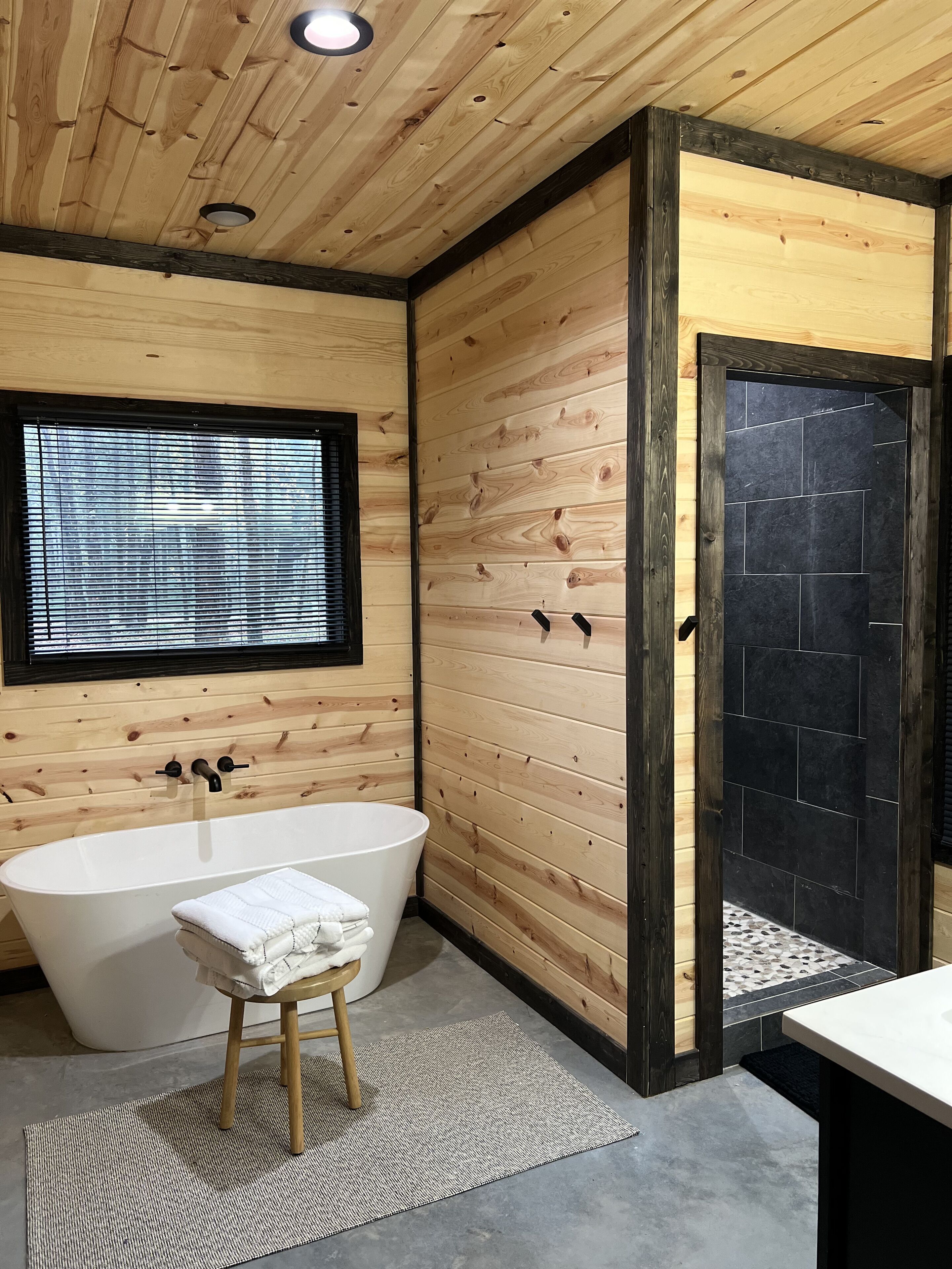 A modern bathroom with a freestanding bathtub, wooden panel walls, a window with blinds, and a black tiled shower area.