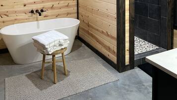 A modern bathroom with a freestanding bathtub, wooden panel walls, a window with blinds, and a black tiled shower area.