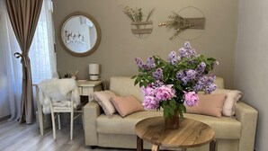 A living room with a beige sofa, a round wooden coffee table, and a vase of flowers. There is a chair, a lamp, and wall decorations.