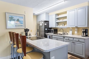 Une cuisine dotée d’un îlot central, de meubles blancs, d’un réfrigérateur en acier inoxydable, d’un four à micro-ondes et d’une machine à café.