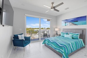 A bedroom with a bed, a chair, a ceiling fan, a TV, and a balcony with a view.