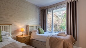 A bedroom with two beds, a nightstand with a lamp, a window with curtains, and a view of trees outside. - Skylark Cottage -  a single-level, two-bedroom property on the gentle slopes of Wansfell Pike, yet o (Ambleside)