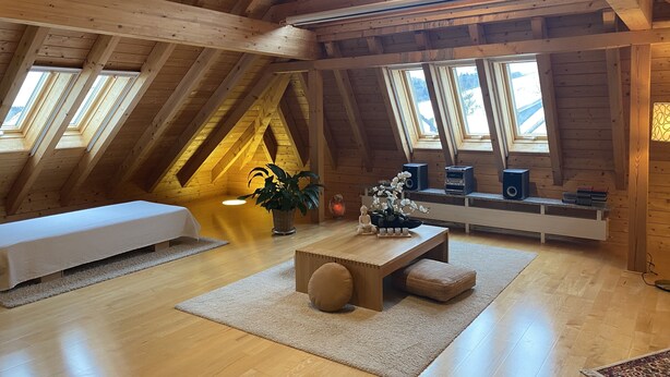 A spacious attic room with wooden beams, a white sofa, a wooden table, and a large window offering a view of the outside.