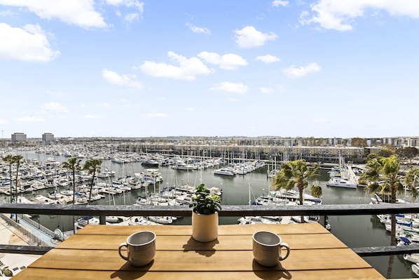A balcony with a wooden table, two mugs, and a potted plant overlooking a marina with numerous boats and a cityscape in the background.