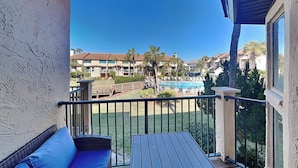 A balcony with a blue sofa and a wooden table, overlooking a swimming pool and a landscaped area with palm trees. - Spyglass 32 by Distinctive Beach Rentals (St. Augustine)