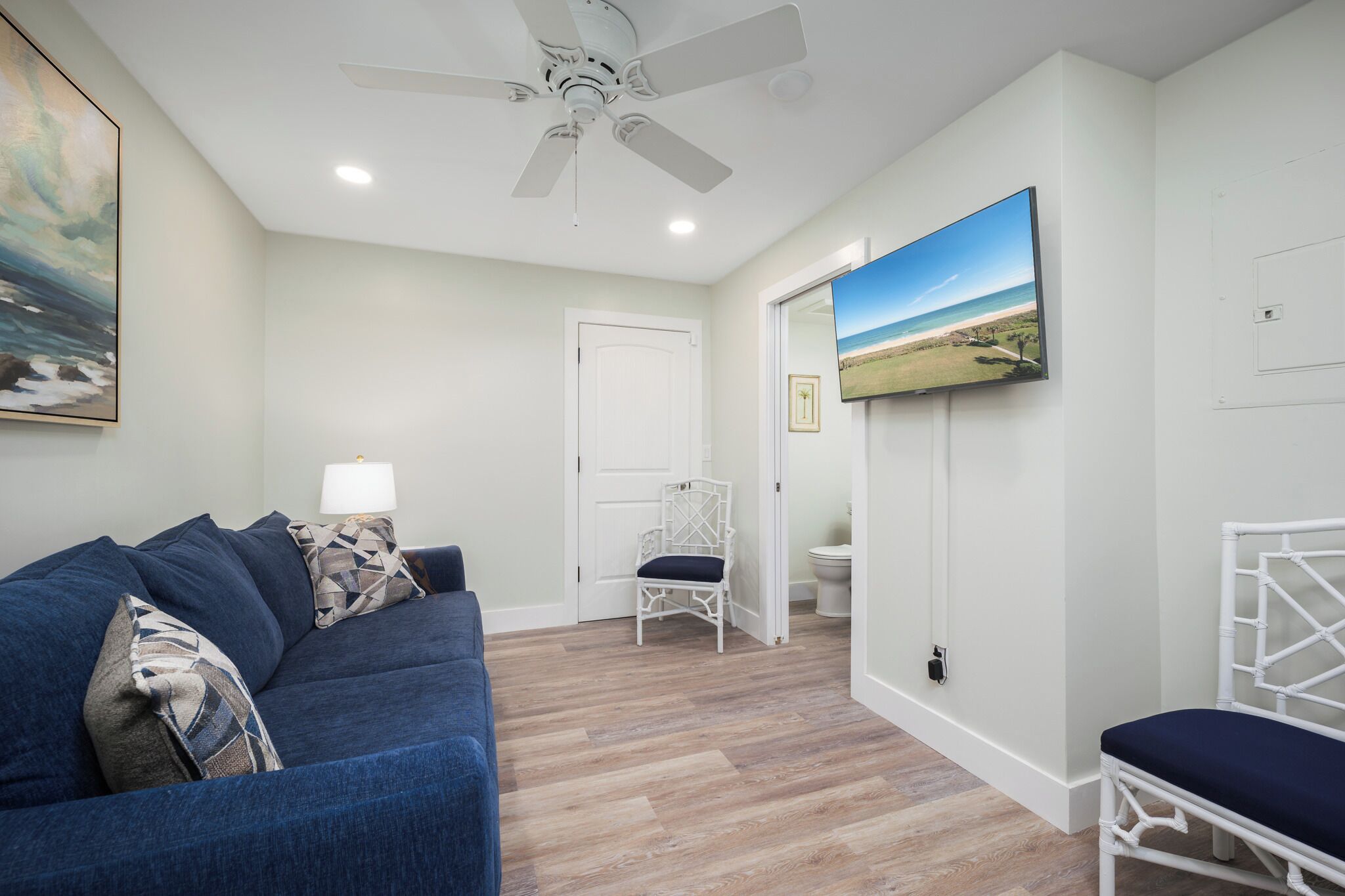 A living room with a blue sofa, white wicker chairs, a ceiling fan, and a painting of a beach.