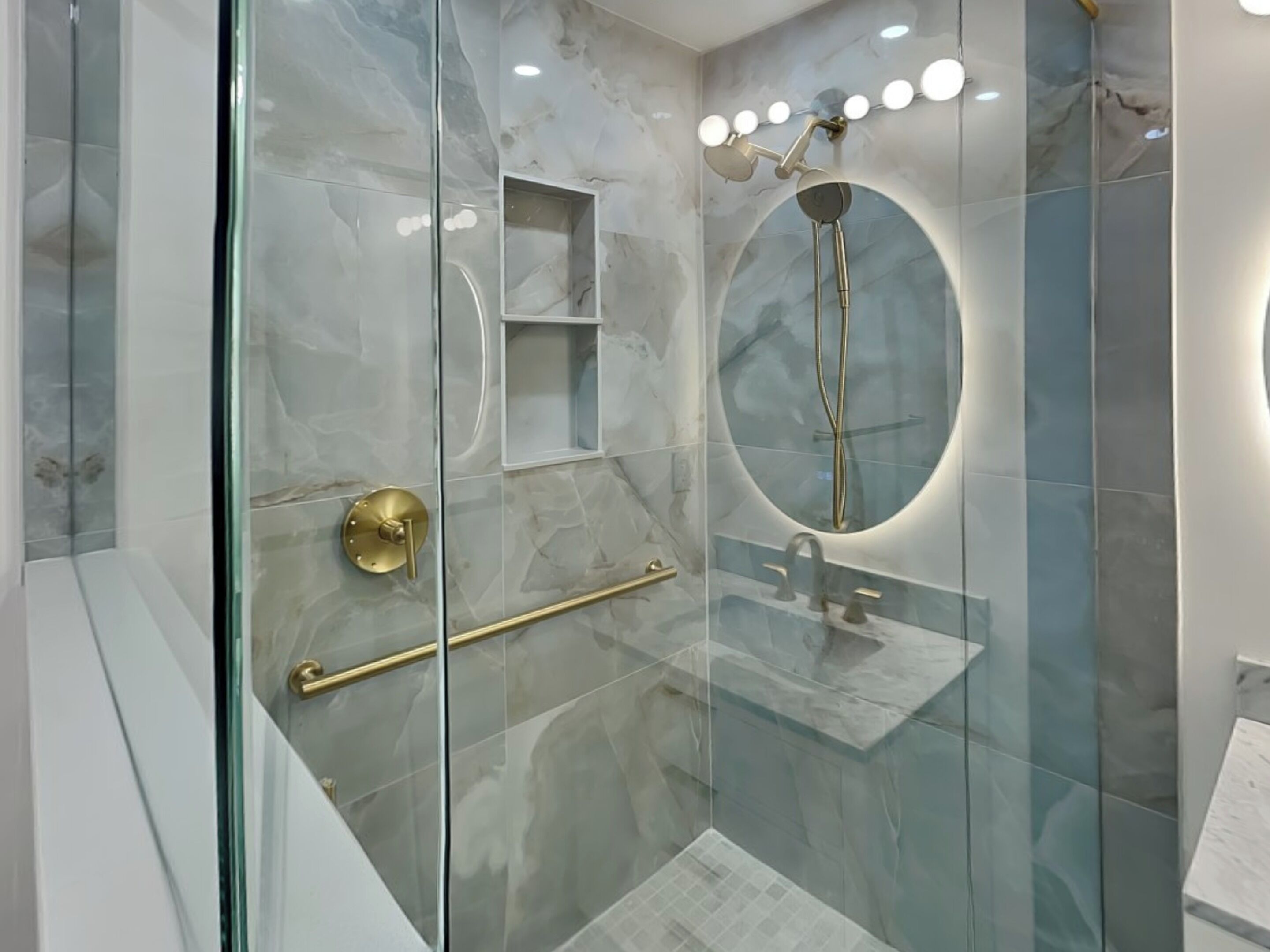 A modern bathroom with a glass-enclosed shower, marble tiles, and a vanity with a round mirror.
