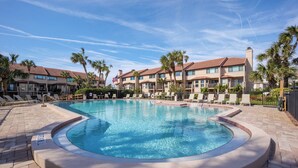 A swimming pool surrounded by lounge chairs and palm trees, with a building featuring balconies in the background. - Spyglass 32 by Distinctive Beach Rentals (St. Augustine)