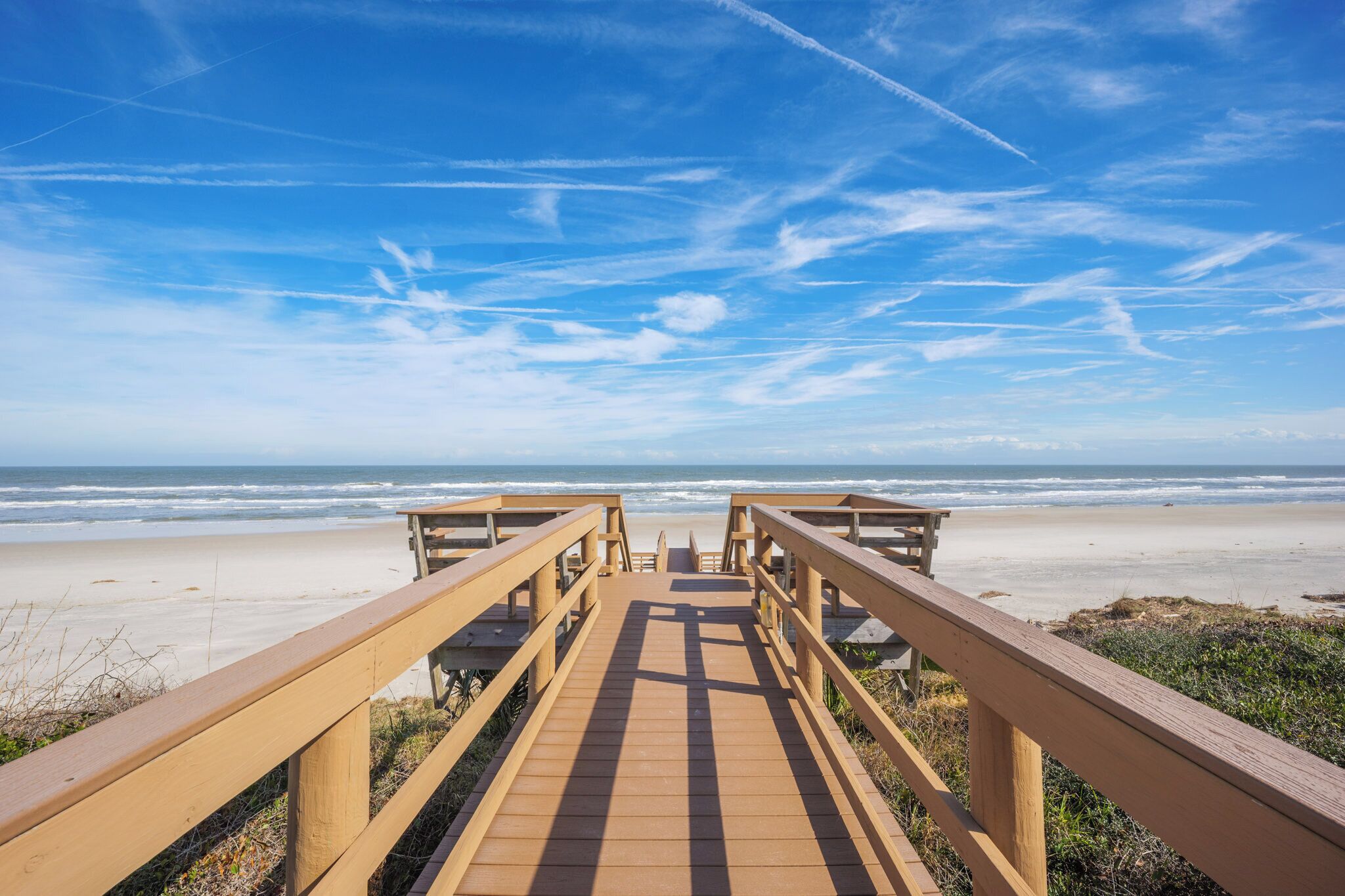 Une promenade en bois qui mène à une plage, avec un ciel bleu dégagé et l’océan en arrière-plan.