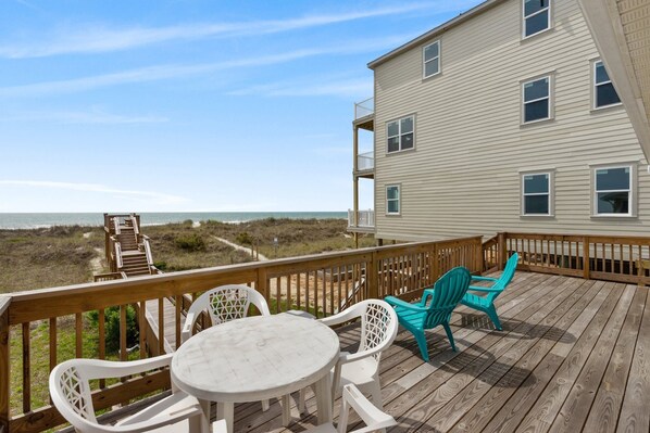 A wooden deck with a table and chairs, overlooking a beach and a building with multiple windows.