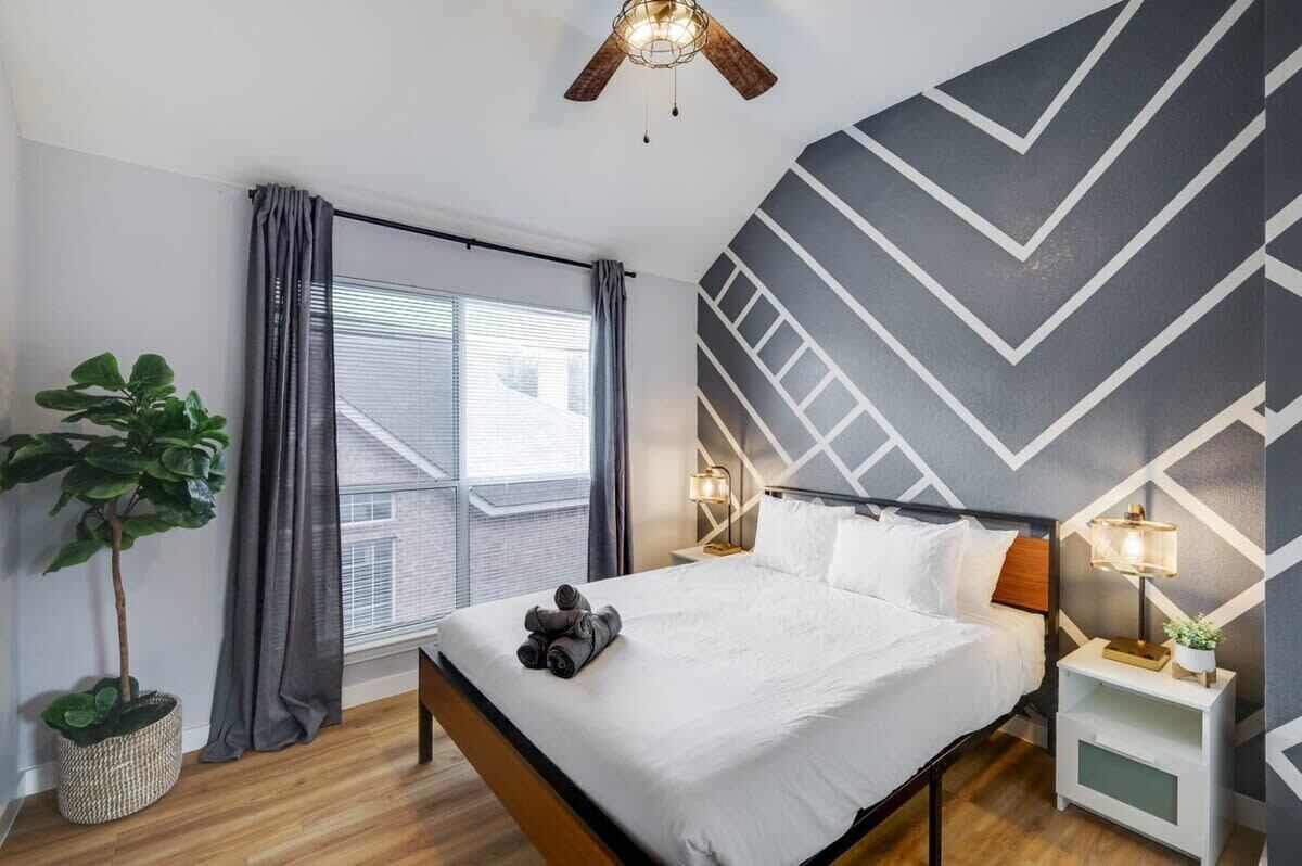 Une chambre d’hôtel moderne, dotée d’un lit, d’une plante en pot, d’une fenêtre avec des rideaux, d’un ventilateur de plafond et d’un motif géométrique sur le mur.
