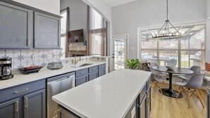 Une cuisine moderne dotée d’un grand îlot central, d’un coin repas avec une table ronde et des chaises, et offrant une vue sur le jardin à travers de grandes fenêtres.