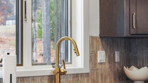 Una cucina moderna con un rubinetto dorato, una finestra vista alberi e uno schienale in legno.