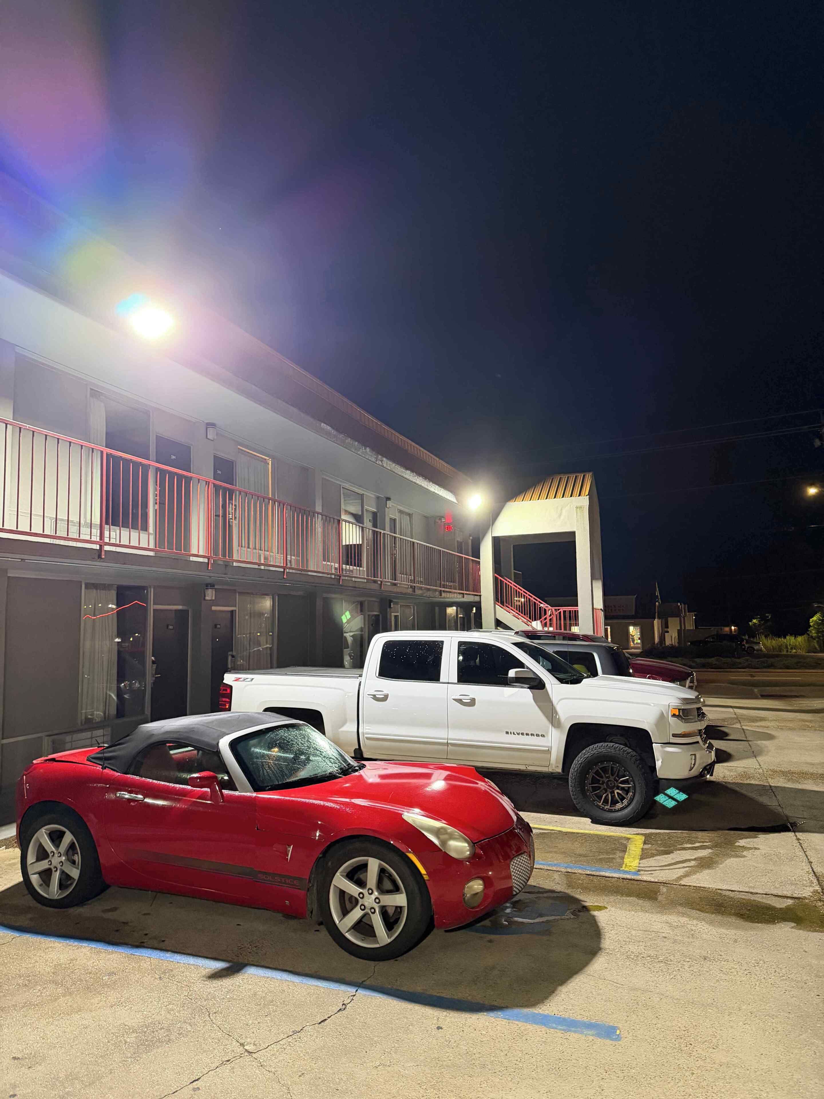 A parking lot with a building in the background, a red convertible, and a white pickup truck.
