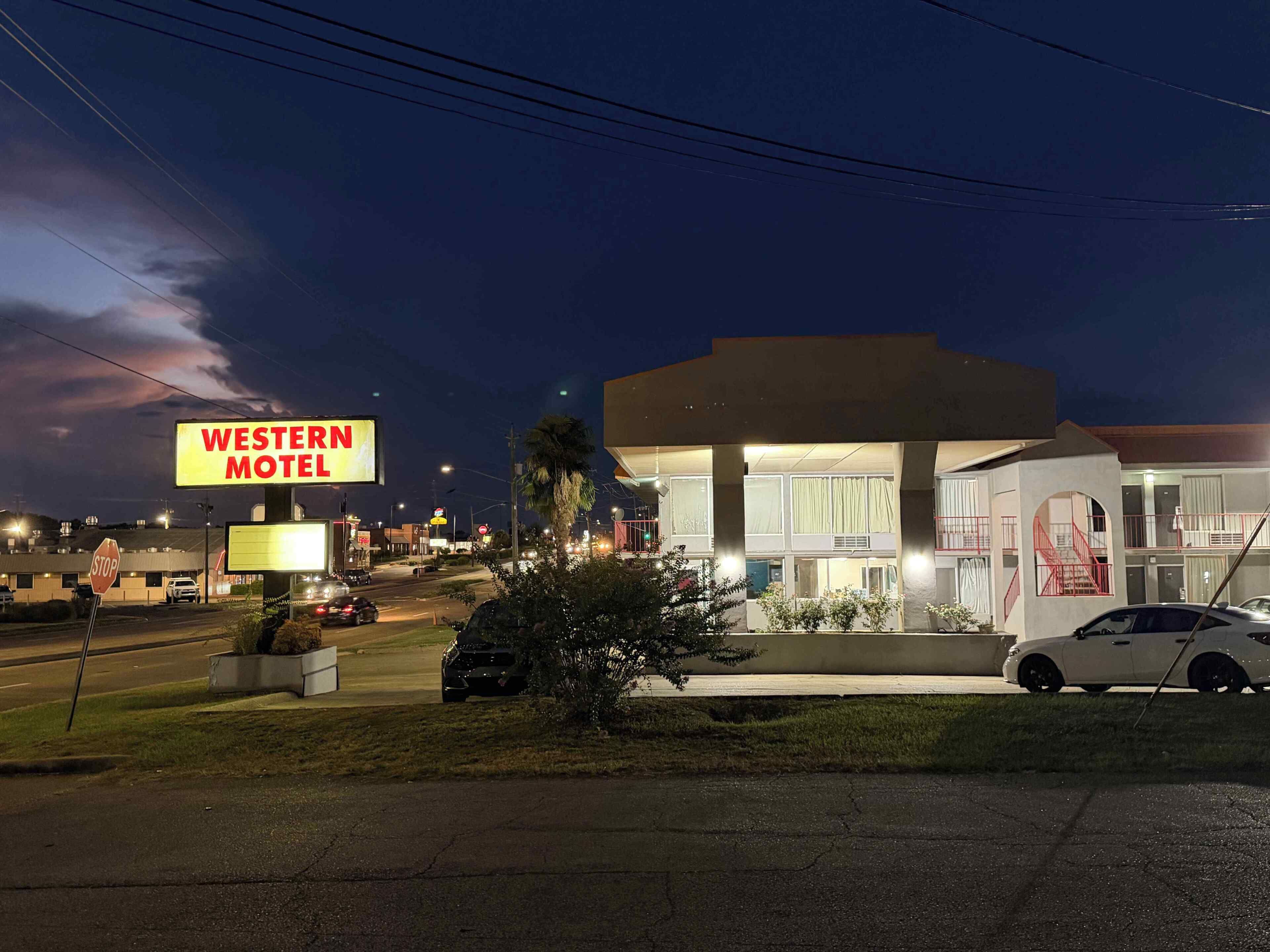 A Western Motel with a large illuminated sign, a stop sign, and a parking lot with cars.