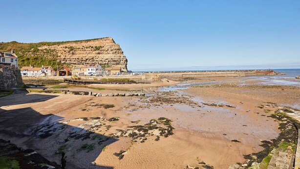 A coastal area with a sandy beach, a cliff, and buildings in the background. - Host & Stay | Sea Haven (Staithes)