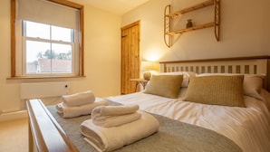 A hotel room with a large bed, a bedside table with towels, a window, and a wooden door. - Maurice Cottage, Docking, Norfolk (Docking)