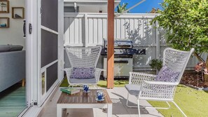 Een terras met twee witte rieten stoelen, een houten tafel en een barbecue.