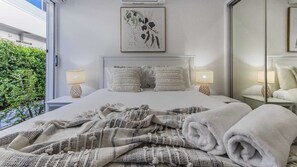 A neatly made bed with two pillows, a bedside lamp, and a framed botanical print on the wall. - Sea Poppy (Casuarina)