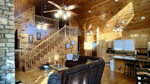 A spacious log cabin interior with a wooden staircase, a living area with a sofa, a kitchen with a bar, and a ceiling fan.