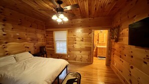 A wooden cabin bedroom with a bed, a ceiling fan, a window, and a view into the kitchen.
