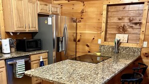 A kitchen with wooden cabinets, a stainless steel refrigerator, a microwave, granite countertops, and bar stools.