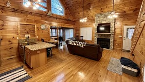 A log cabin interior with a kitchen, dining area, and living room, featuring wooden floors, a stone fireplace, and large windows.