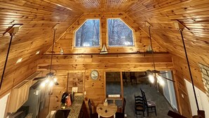 An attic room with wooden slats on the ceiling, two windows, a clock, and a table with chairs.