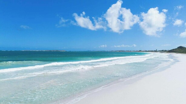 Een strand met helder turquoise water, wit zand en een verre kustlijn met gebouwen.