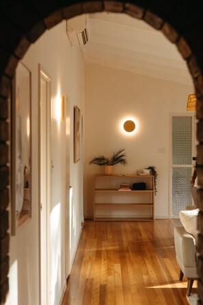 A hallway with wooden flooring, a white wall, a round light fixture, and a bookshelf with plants.