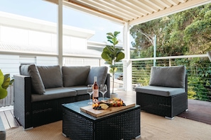 A covered outdoor seating area with wicker furniture, a wooden table with fruit and wine, and a view of greenery.