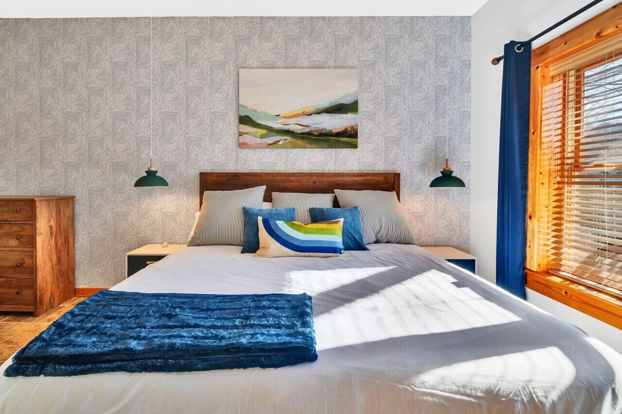 Een slaapkamer met een groot bed, twee nachtkastjes, een houten hoofdeinde, een schilderij aan de muur en een raam met jaloezieën.