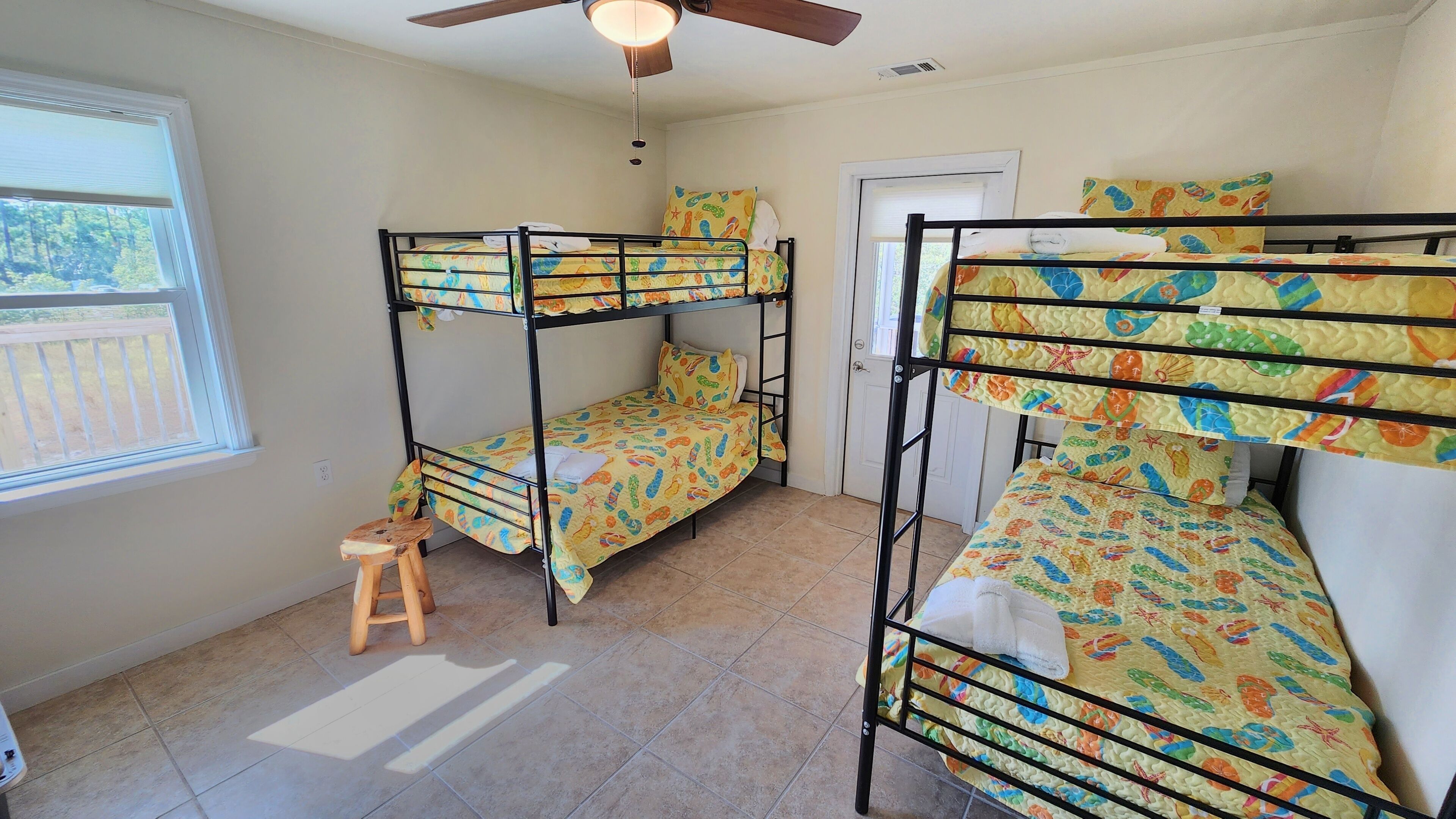 A dormitory room with bunk beds, a ceiling fan, a window, and a small stool.