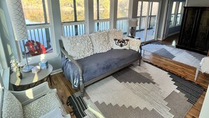 Een gezellige woonkamer met een bank, een bijzettafeltje en een fauteuil. Grote ramen bieden uitzicht op bomen en een meer.