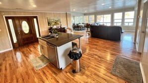 Een ruime woonkamer met een keukeneiland, houten vloeren en grote ramen die veel natuurlijk licht binnenlaten.