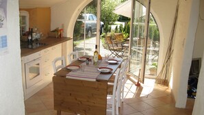 Un coin repas avec une table en bois dressée pour quatre personnes, des chaises blanches et une vue sur l’extérieur grâce à de grandes fenêtres.